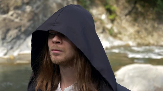 Close-up Shot. Man In Black Cloak With Hood Cowl Walking On Rocks Alongside Mountain River In The Wood. Summer Sunny Day. 4K Slow Motion