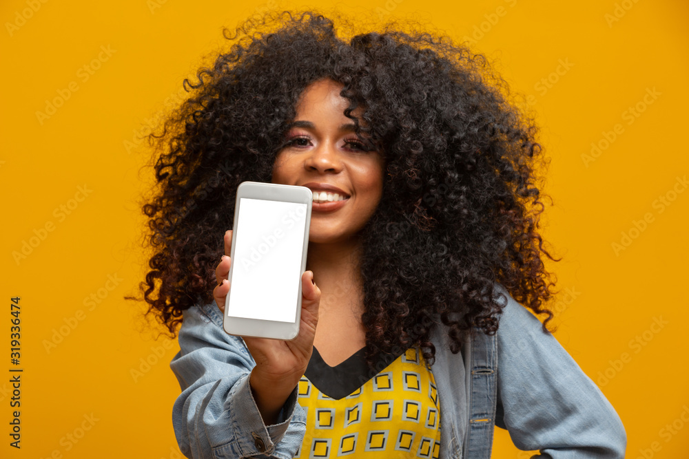 Wall mural Portrait of cheerful, positive, attractive young woman in jeans shirt, having smart phone with white screen in hand, pointing with forefinger to product, isolated on yellow background