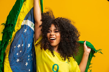 Afro girl cheering for favorite brazilian team, holding national flag in yellow background. - obrazy, fototapety, plakaty
