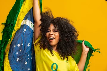 Fotobehang Afro girl cheering for favorite brazilian team, holding national flag in yellow background. © Brastock Images