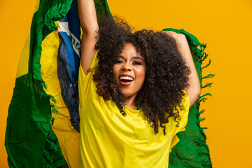Afro girl cheering for favorite brazilian team, holding national flag in yellow background.