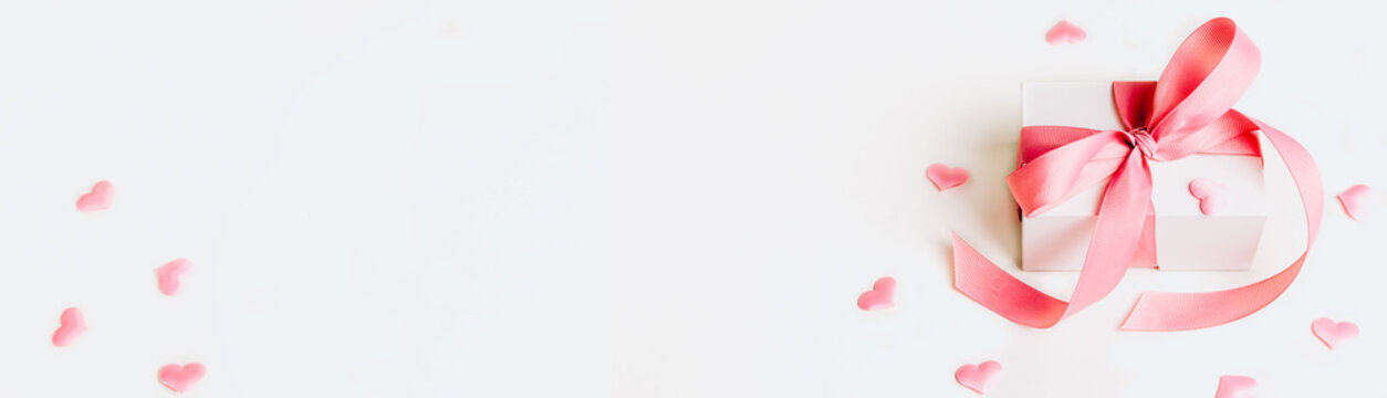 White gift box with pink ribbon and a small pink hearts on white background. Selective focus. Web banner format