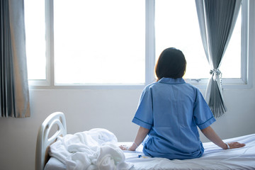 Back view of patient woman sitting on bed in hospital ward, looking away at window and hope...