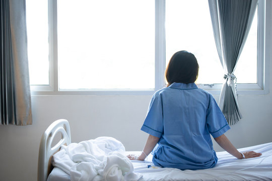 Back view of patient woman sitting on bed in hospital ward, looking away at window and hope everything will be better. Conceptual of patien lifestyle in hospital.