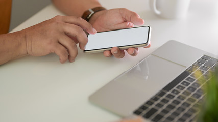 Cropped shot of male freelancer texting on smartphone in workspace with laptop on white table