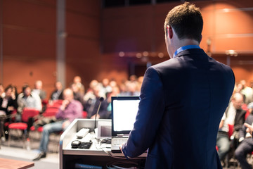 Speaker giving a talk on corporate business conference. Unrecognizable people in audience at...