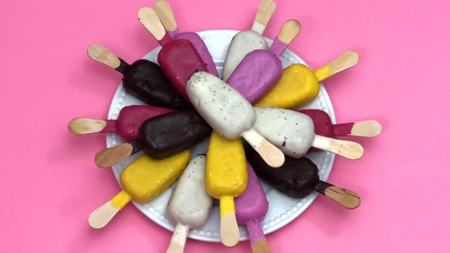 HD video of Multiple colorful colored candy coated popsicle cake pops on an off white porcelain plate rotating on pink background with colorful paper confetti. Zooming out. Fun food for kids parties.