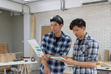 Two young asian carpenters holding paint color sample chart and notebook in workplace - Handyman and diy concept