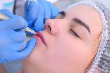 Cosmetologist making lips microblading procedure for girl using tattoo machine, face closeup, side view. Beautician is applying permanent makeup on woman's lips. Beauty industry concept.