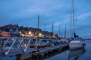 View of the Volendam harbor and yacht, boats and sailing boats anchored, Volendam, Netherlands. Marine with illuminated houses and blue sky.