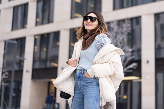 European model in a beige oversized down jacket, knitted sweater with a handbag and glasses posing near the office building. Lifestyle. Real emotions