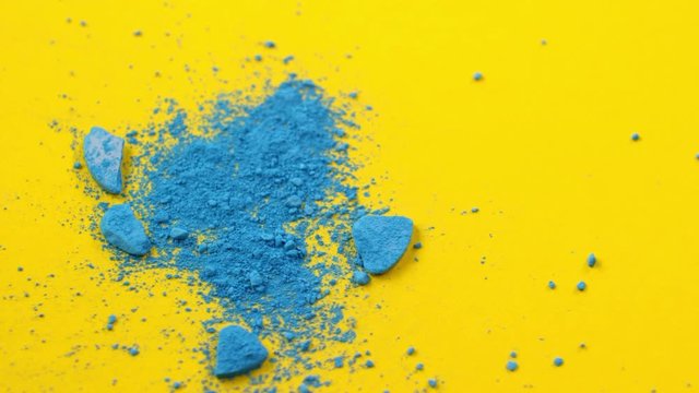 Close-up tracking shot of crushed blue eyeshadow powder being blown off table with bright yellow surface