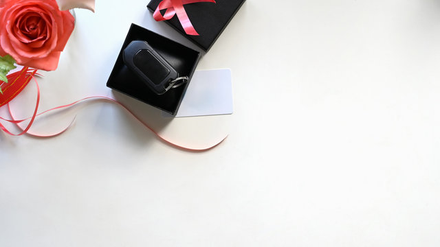 Top view Digital car key putting inside the black gift box with red ribbon,bouquet of roses and wish card on the white desk as background. Surprising Valentine's Day gift concept.