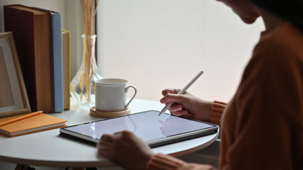 Cropped shot side view of young creative woman drawing/using on a white blank screen computer tablet at her modern desk. Books, coffee cup, pencil, wild grass in jug all of these putting on desk.