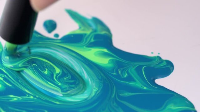 Close up tracking shot of female hand stirring green and blue nail polishes together with brush and making beautiful bright mixture on table with white surface