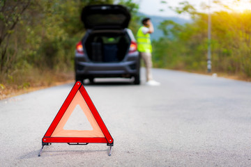 Breakdown triangle sign on road with worried Asian man talking by mobile phone with insurance or car service center company after car breakdown on country road in background..
