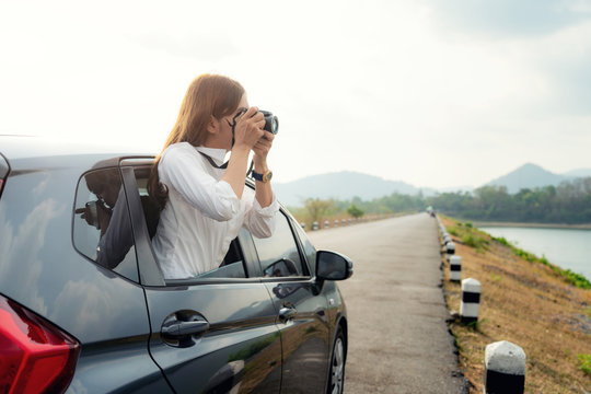 Young Asian Woman tourist taking photo in car with camera driving on road trip travel vacation. Girl passenger taking picture out of window with beautiful view lake and mountains in background..