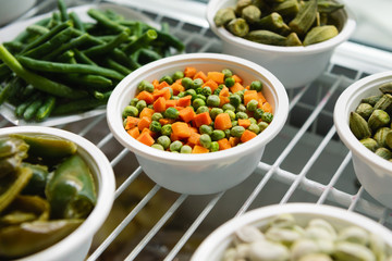 A frozen mixture of carrots and green peas lies on a white plate. Next to the plates are other frozen vegetables and fruits.