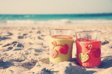 Two cups in knitted covers stand on a sandy beach in the summer.