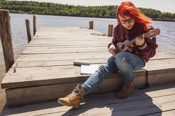 Caucasian teenager girl in glasses with bright red hair plays ukulele