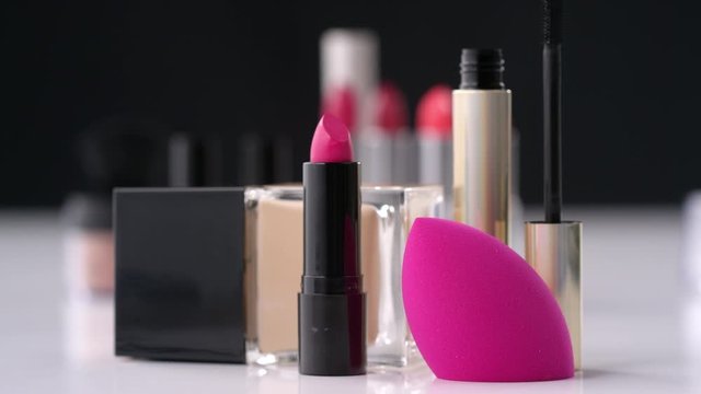 Tracking close up shot of pink lipstick, foundation, mascara and makeup sponge on table with decorative cosmetics