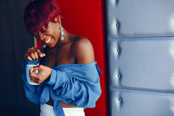 beautiful and stylish dark-skinned girl with red short hair, dressed in a blue top with long sleeves and white pants standing in a cafe and drinking a coffee