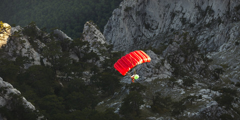 Base jumper under a bright red parachute flies against a background of a dark rocky landscape after jumping from the top of a mountain, view from above. Panorama.