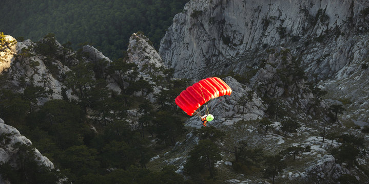 Base Jumper Under A Bright Red Parachute Flies Against A Background Of A Dark Rocky Landscape After Jumping From The Top Of A Mountain, View From Above. Panorama.