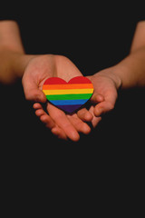 Female arm holding the heart coloured in LGBT pride colours on the dark background. Concept of the International Day Against Homophobia concept, sexual equality, feminism, social safety