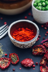 A cup of red spice made of red peppers on a dark table top surrounded with dehydrated small tomatoes