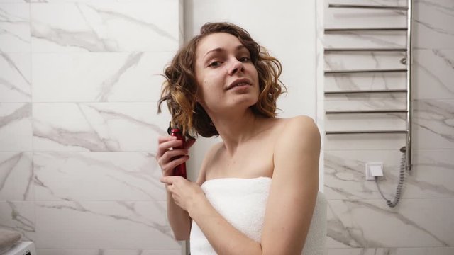 Smiling young woman in big white bath towel styling her hair with a curling iron in front of a mirror. A girl after shower makes a hairstyle in the bathroom. Front view. Marble wall on background