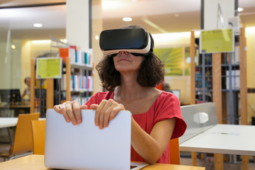 Focused user with virtual reality glasses holding laptop screen. Thoughtful Caucasian woman with VR headset sitting at library. Technology concept