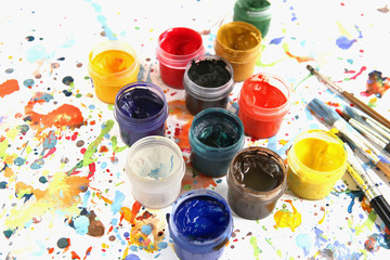 Jars with gouache paints and brushes on colorful paint splashes background. Plastic cans with multi-colored dye for drawing..