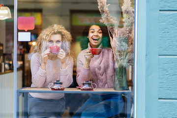 Two multi ethnic friends enjoying coffee together in a coffee shop viewed through glass with...