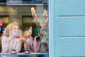Two multi ethnic friends enjoying coffee together in a coffee shop viewed through glass with reflections as they sit at a table chatting