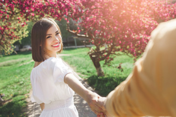 Woman holding her boyfriend by the hand. Young couple together walking in the garden. Lovers outdoors.