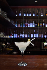 White cocktail on bar counter. Drink with martini, gin, tequila and lime on a background of bottles with alcohol. Glass with booze on a wooden board. Blue background with ultraviolet illumination.