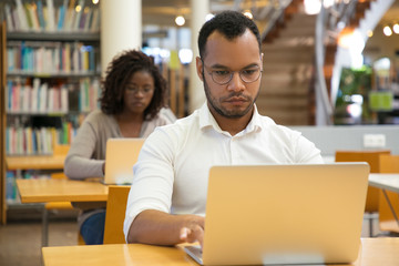 Concentrated man typing on laptop. Handsome young student sitting at library and using modern device. Technology concept