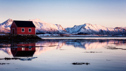 Obraz premium Amazing winter landscape with red rorbu, traditional scandinavian fishing house, snowy mountains in sunset light, blue sky and reflection in water, Lofoten Islands, Norway. Outdoor travel background