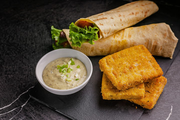 Close up tortilla wrap with salmon and fresh salad, breaded fish fillet and sauce on a black...