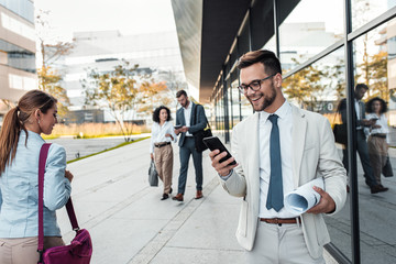 Portrait of smiling businessman standing in front of modern office buildings looking at phone.