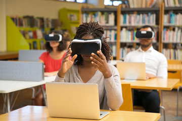 Concentrated young woman with virtual reality glasses. Focused woman sitting in library and...