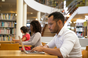 Side view of focused man typing on laptop. Handsome young student sitting at library and using modern device. Technology concept