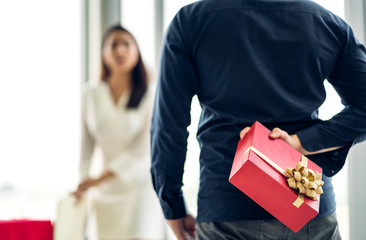 Young handsome man giving gift box surprise to girlfriend and holding gift behind back at home