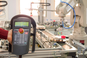 Close up of man's hand holding teach panel to control robot arm which is integrated on smart factory production line. industry 4.0 automation line equipped with sensors and robot at background.