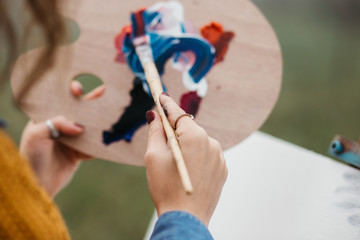 Close up photo of young female artist working on painting outdoors. She holds oil paints, artist...
