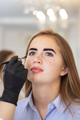 A female cosmetologist performs eyebrow correction on a beautiful model in a beauty salon. Applying freckles to the face, skin care and salon treatments. Model with vitiligo pigmentation. 