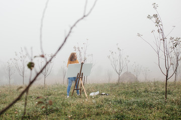 Young female artist working on painting outdoors. She is in front of the canvas and drawing.She holds oil paints, artist brushes, canvas and palette. Portrait of concentrated woman with curly hair. 