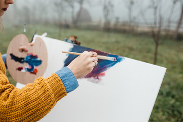 Close up photo of young female artist working on painting outdoors. She holds oil paints, artist brushes, canvas and palette. She is mixing colours on palette. Hands close up.