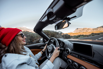 Happy woman in red hat driving convertible car while traveling on the desert road. Carefree...